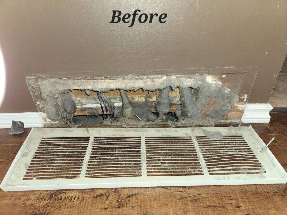 Floor-level wall return opening choked with thick hanging dust, the dusty grille lying on the hardwood floor