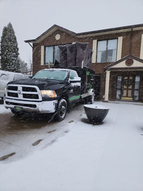 The black vacuum truck in a snowy driveway with headlights on during a snowfall